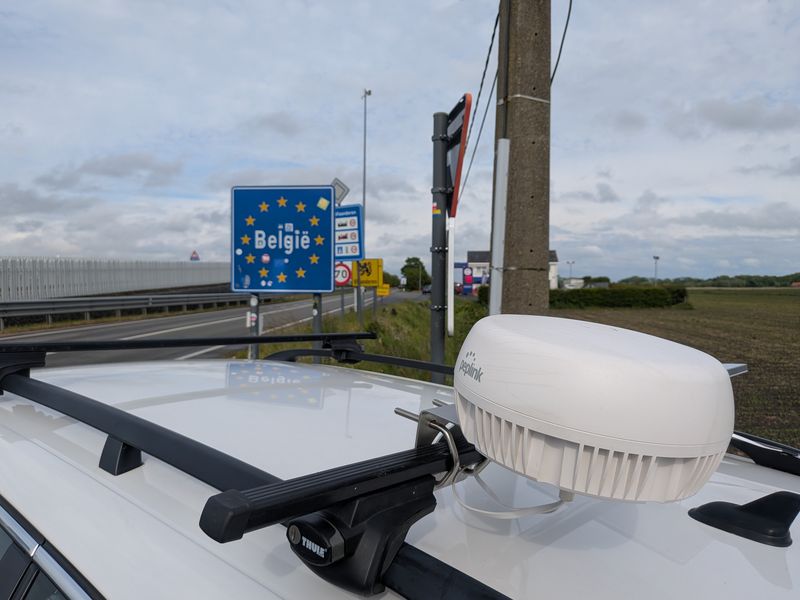 A bonded antenna dome on the trial car's roof rack, with the België border sign behind.